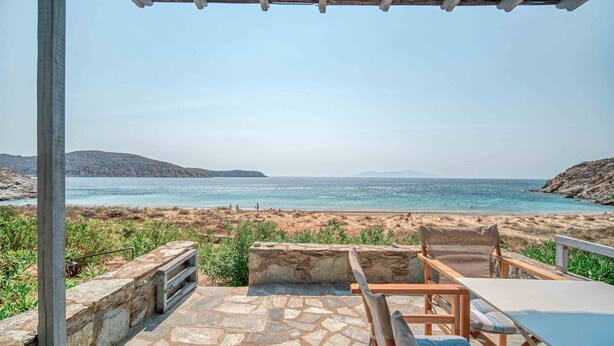 A patio with stone flooring, a table, and two chairs overlooking a beach with a rocky shoreline and distant hills. - Holiday Home 'Karavi' with Private Terrace, Balcony and Air Conditioning (S???f??)
