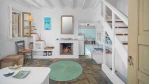 A room with a stone floor, white walls, a fireplace, a dining area with a table and chairs, and a kitchen visible through an open door. - Holiday Home 'Karavi' with Private Terrace, Balcony and Air Conditioning (S???f??)