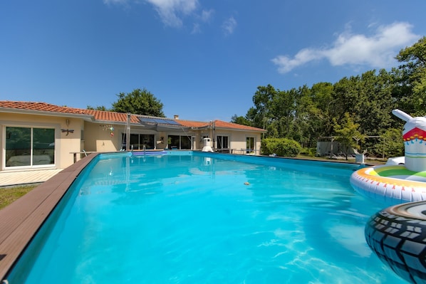 A swimming pool with a wooden deck, a house with a tiled roof, and trees in the background.