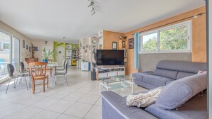 A living room with a sofa, TV, dining table, chairs, and a kitchen area in the background.