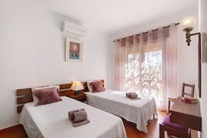 A room with two beds, a table, a chair, an air conditioning unit, and a window with sheer curtains.