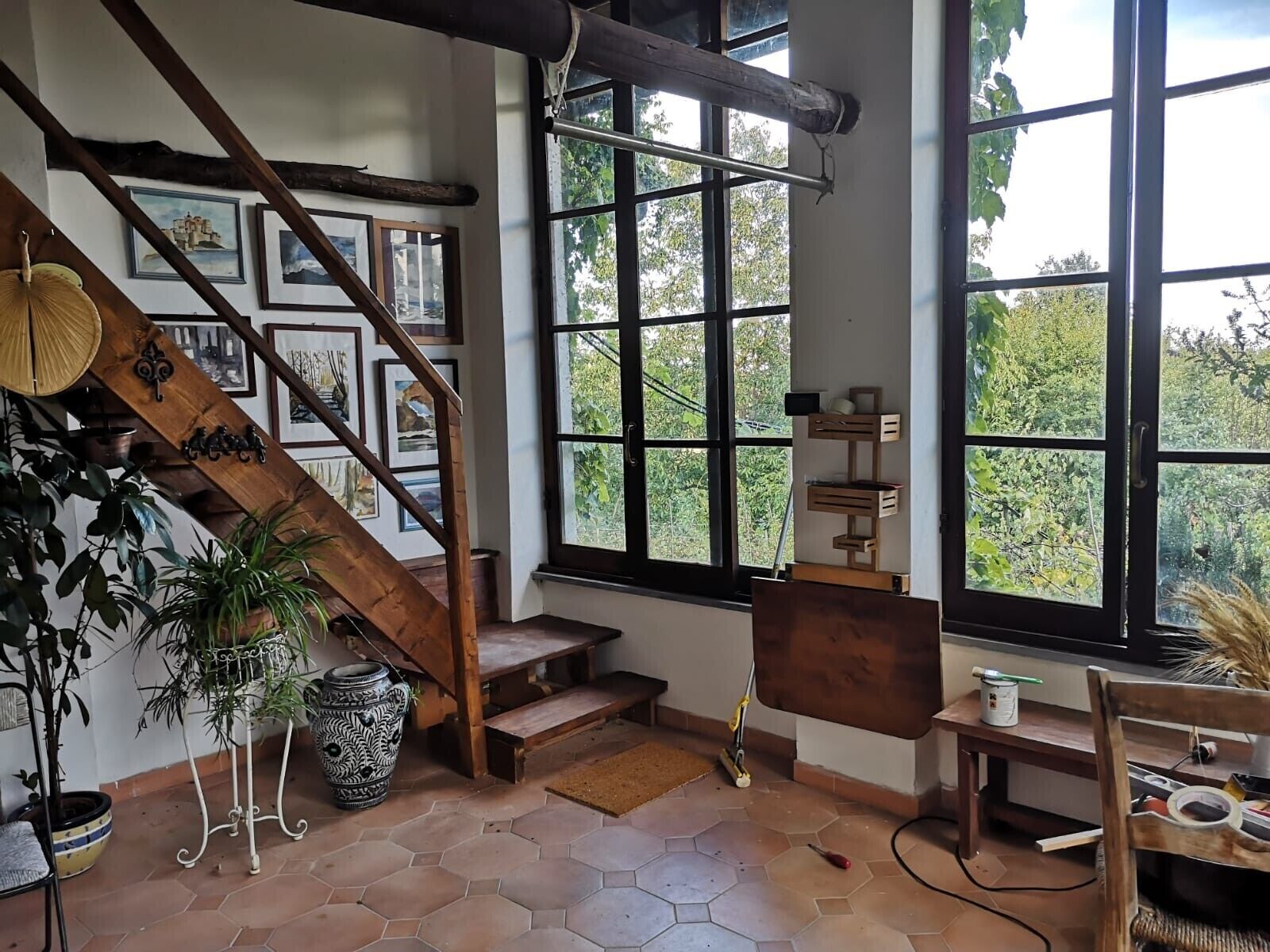 A room with a wooden staircase, large windows, and a view of greenery outside.