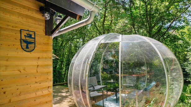 A transparent dome structure with a table and chairs inside, adjacent to a wooden building with a coat of arms. - Cottage 'Gîte La Marinière' with Private Terrace and Private Garden (Plurien)