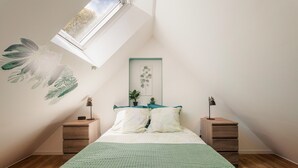 Un dormitorio con techo inclinado, una cama con cubrecama verde, dos mesitas de noche con lámparas y una planta colgada en la pared.