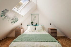 Un dormitorio con techo inclinado, una cama con cubrecama verde, dos mesitas de noche con lámparas y una planta colgada en la pared.
