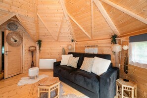 A log cabin interior with a dark sofa, wooden walls, a ceiling clock, and a rustic decor style. - Chalet 'Gite Petite Chaumière Kota Finland' with Private Terrace, Garden and Air Conditioning (Plurien)