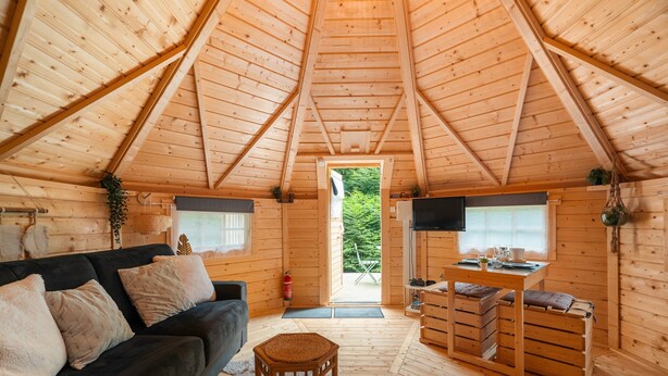 Ein gemütliches Blockhaus-Interieur mit Holzbalkendecke, einer Couch, einem Esstisch und Blick durch eine Tür.