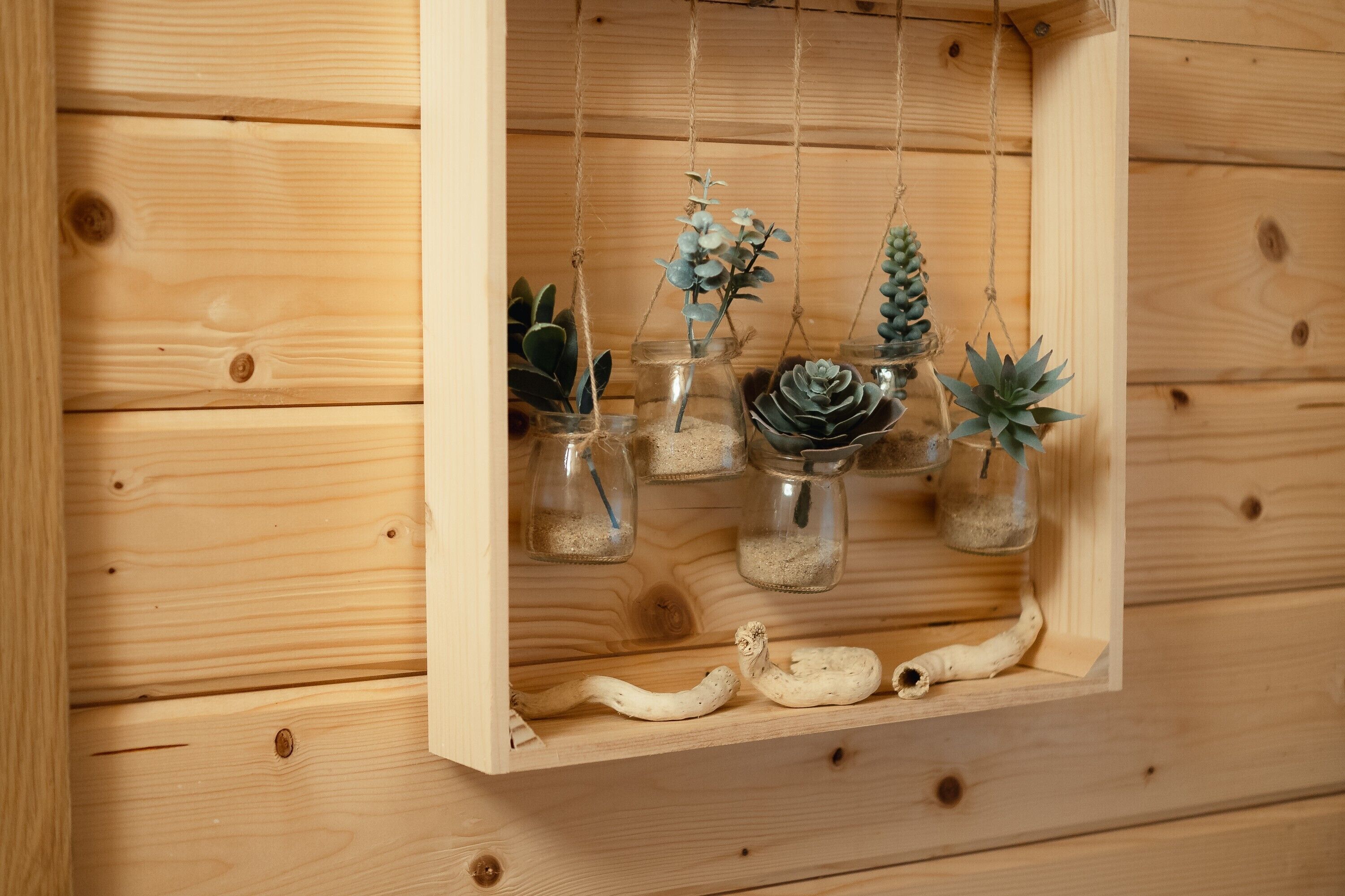 Exhibición de madera con frascos de vidrio colgantes que contienen plantas y un objeto blanco.