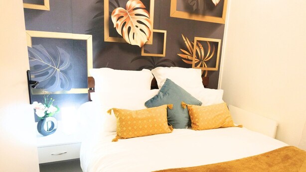 Ein ordentlich bezogenes Bett mit einer braunen Decke, ein Nachttisch mit einer Lampe und eine Wand mit gerahmten Kunstwerken.
