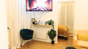 Ein Wohnzimmer mit an der Wand montiertem Fernseher, einem blauen Sessel, einem Bett mit brauner Decke und einem gelben Sofa.