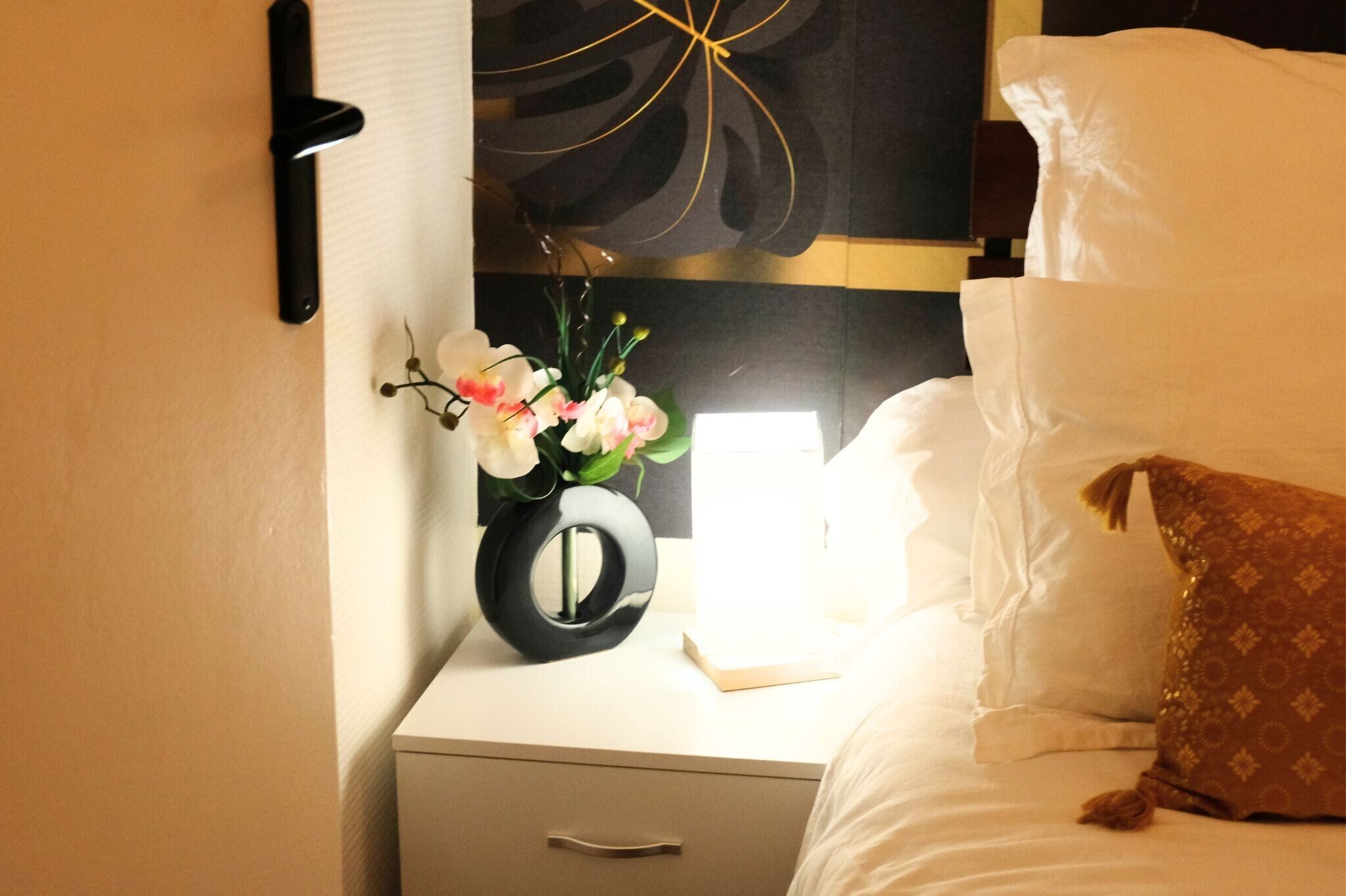 A neatly made bed with white linens, a brown decorative pillow, and a bedside table with a black vase containing flowers.