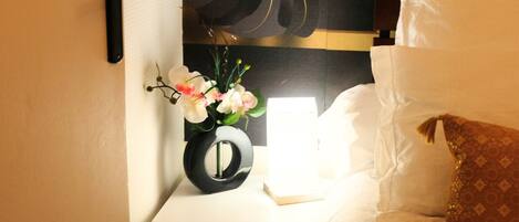 A neatly made bed with white linens, a brown decorative pillow, and a bedside table with a black vase containing flowers.