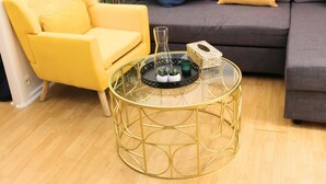 Ein gelber Sessel, ein runder Glastisch mit goldener Rahmen und ein graues Sofa mit gelben Kissen in einem Wohnzimmer.