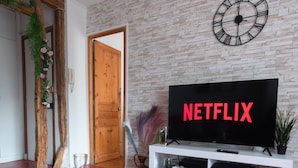 A living room with a wooden coffee table, a white entertainment unit with a flat-screen TV displaying 'NETFLIX,' a clock on the wall, and a red herring-patterned floor.