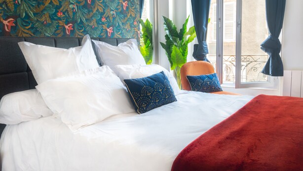 A bed with white and blue pillows, a red blanket, and a floral-patterned wall behind it.
