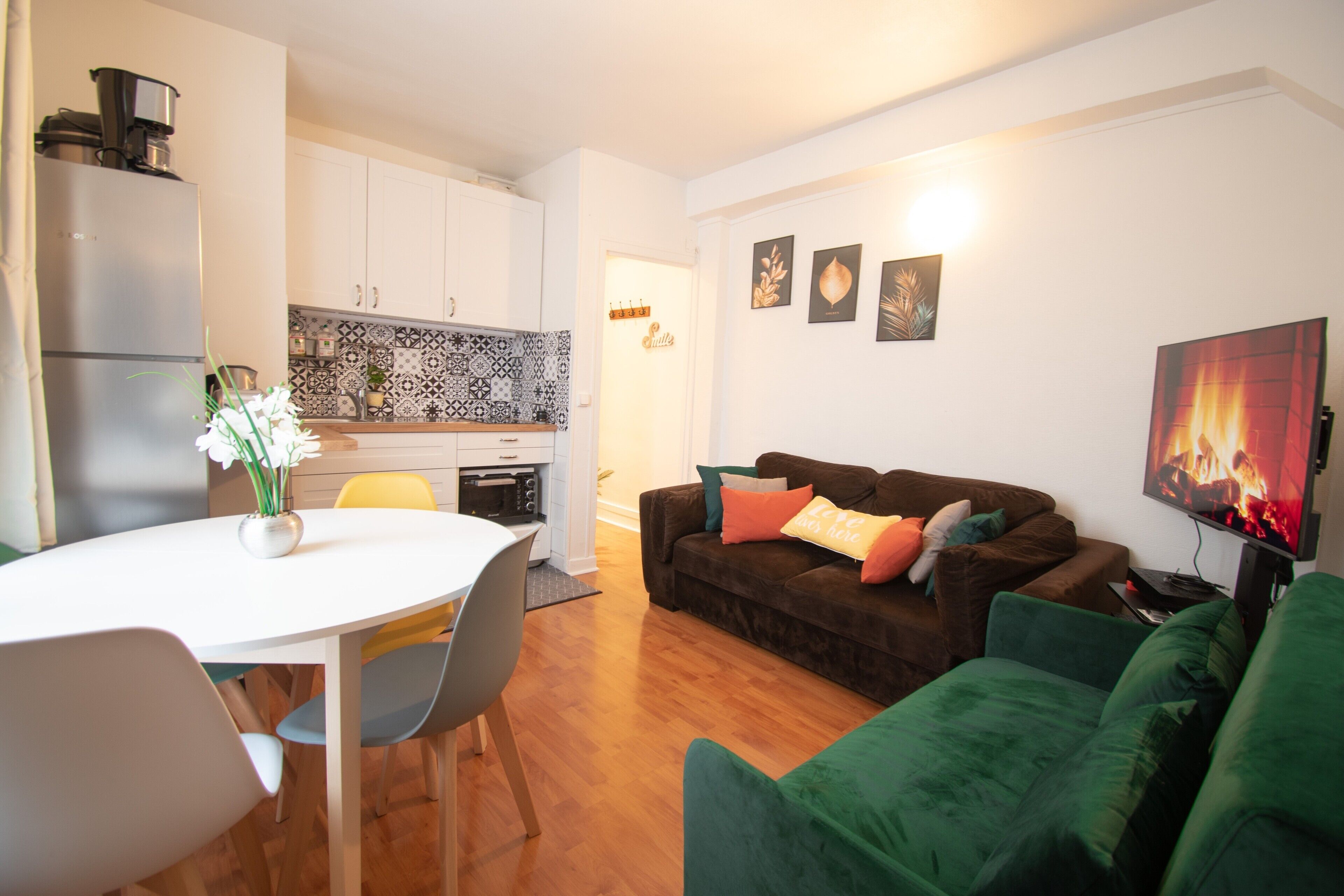 A modern kitchen with white cabinets, a dining table with chairs, a brown sofa with cushions, and a flat-screen TV.