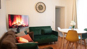 A living room with a green sofa, a TV displaying a fireplace, a dining table with chairs, and a clock on the wall. - Apartment 'Appartement 2 Pièces À Paris' with Wi-Fi (Gentilly)