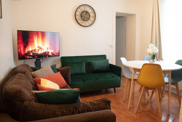 A living room with a green sofa, a TV displaying a fireplace, a dining table with chairs, and a clock on the wall.