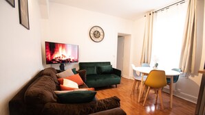 A living room with a sofa, a television displaying a fireplace, a dining area with a table and chairs, and a clock on the wall. - Apartment 'Appartement 2 Pièces À Paris' with Wi-Fi (Gentilly)
