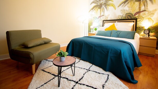 A bedroom with a bed, a chair, a small table with a plant, and a wall with a palm tree mural. - Apartment 'Appartement 2 Pièces À Paris' with Wi-Fi (Gentilly)
