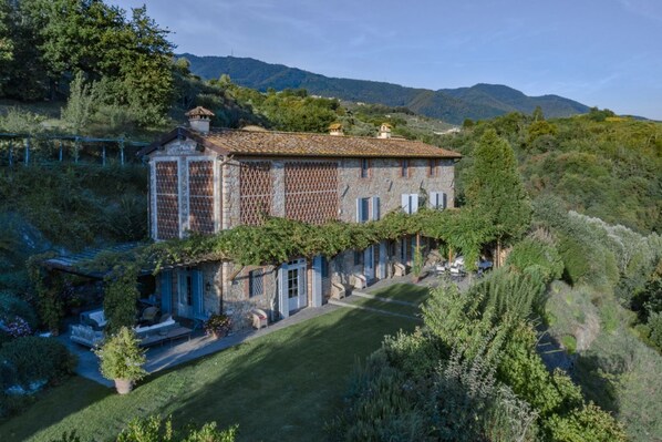 Un bâtiment en pierre, surmonté d’un toit en tuiles, entouré d’une végétation luxuriante et de collines.