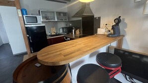 Een keuken met een houten bar, twee krukken, een magnetron, een koffieapparaat en een koelkast.