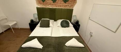En pænt redt seng med grønt dynebetræk og hvide sengetøj, to sengeborde med lamper, en hvid stol, og en murstensvæg med grønne dekorationer af blade.
