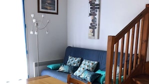 A dining area with a wooden table and a glass bowl centerpiece. A blue sofa with patterned cushions is against the wall, and a staircase is visible to the right. - Charmante Maisonnette à Proximité Immédiate de la Plage (BISCARROSSE PLAGE)
