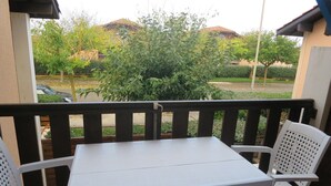 Un balcón con una mesa blanca y dos sillas blancas, con vista a un área verde con árboles y un edificio al fondo.