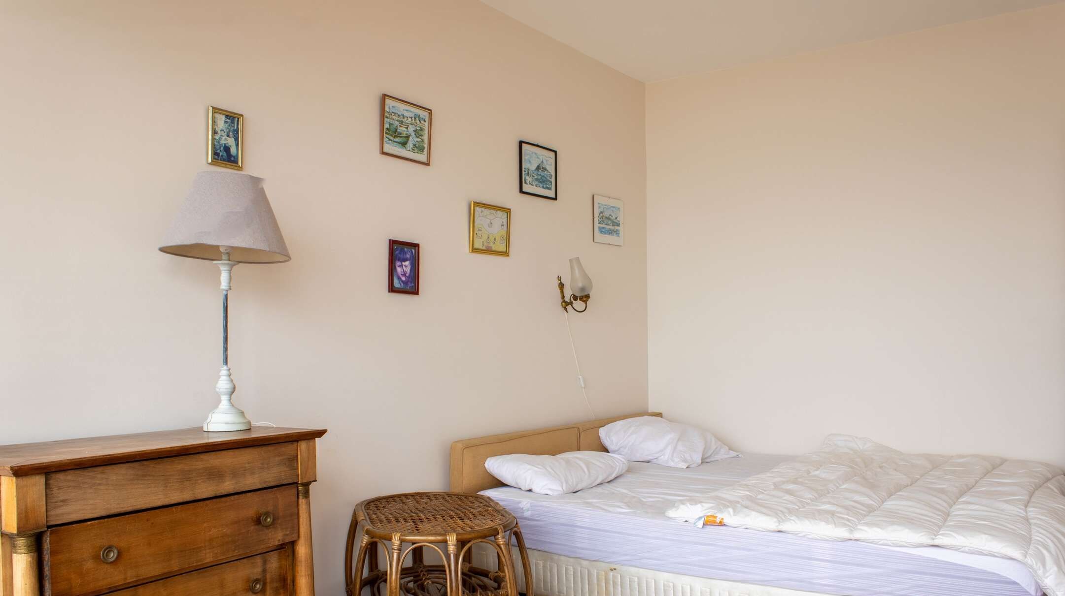 Une chambre à coucher avec un lit, une commode en bois, un tabouret en osier et des tableaux accrochés au mur.