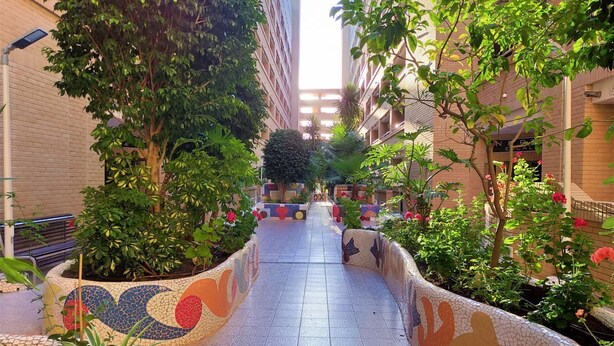 A corridor with tiled flooring, surrounded by greenery and buildings.