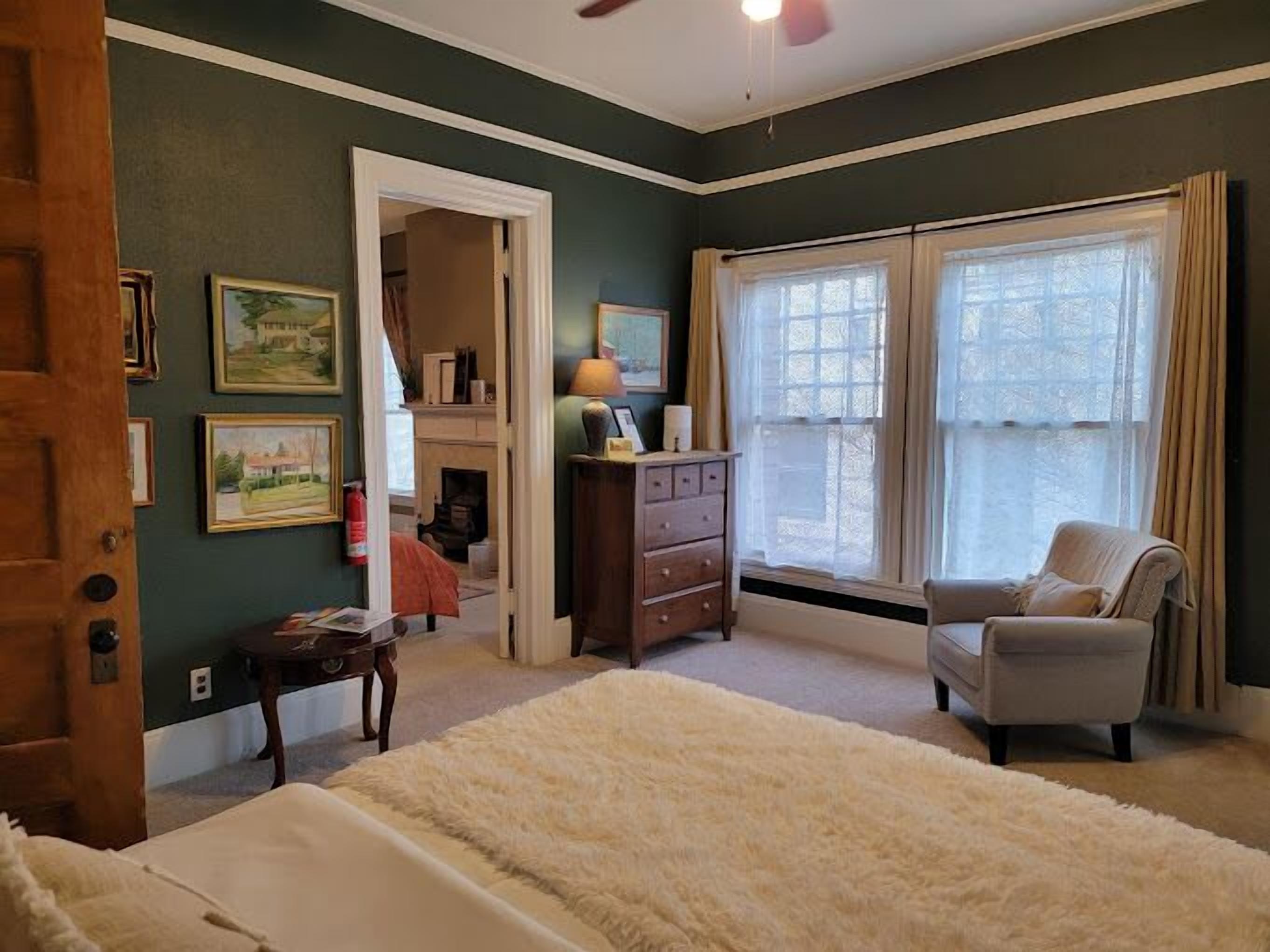 Un dormitorio con cama, una silla, cómoda, espejo y ventana con cortinas.