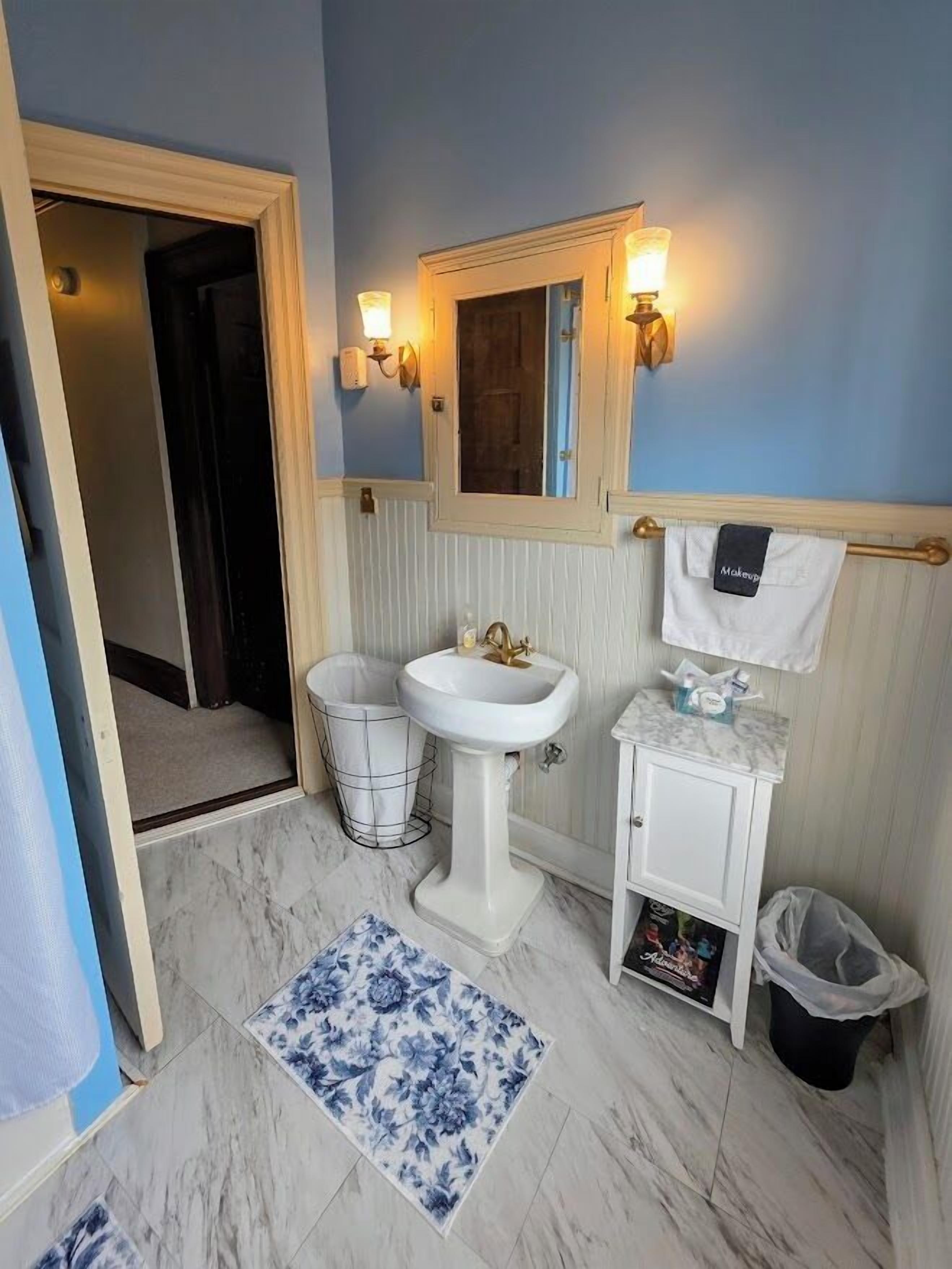 A bathroom with a pedestal sink, vanity, and a mirror. There is a trash can and a small trash bin.