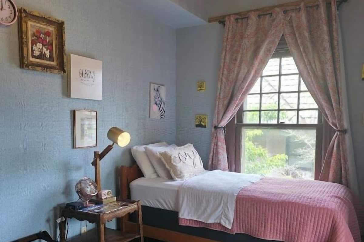 Ein Schlafzimmer mit einem Einzelbett, einem Nachttisch mit Lampe, einem kleinen Stuhl, einem Teppich und einem Fenster mit Vorhängen.