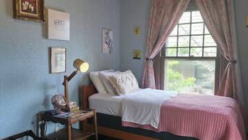 Un dormitorio con una cama individual, una mesita de noche con lámpara, una silla pequeña, una alfombra y una ventana con cortinas.