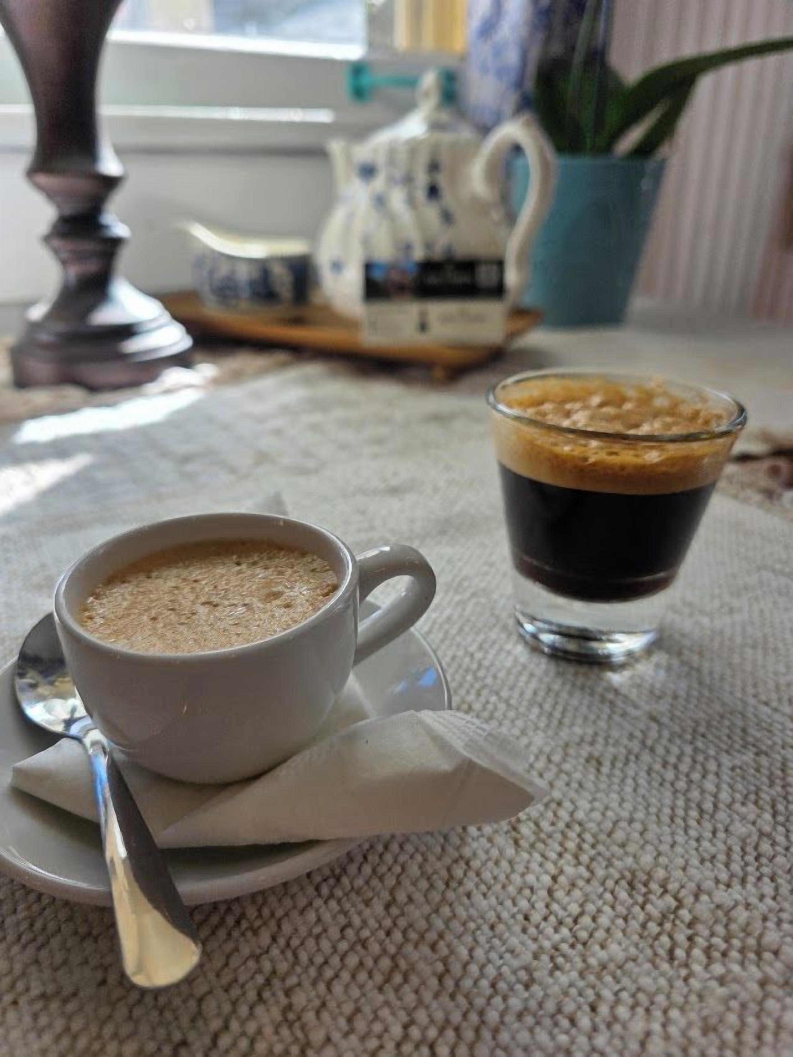 Une tasse de café posée sur une soucoupe, avec une cuillère, et un verre contenant un liquide sombre et mousseux, le tout sur une table.