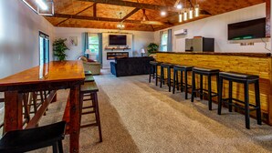 A spacious living area with a wooden ceiling, a large TV mounted on the wall, a fireplace, and a bar area with stools. - New Spacious Warren Dunes Beach & Winery Retreat w/Hot Tub & Pool Table (Bridgman)