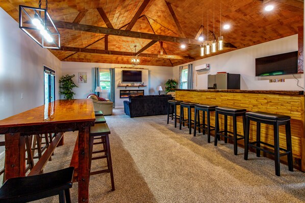 A spacious living area with a wooden ceiling, a large TV mounted on the wall, a fireplace, and a bar area with stools.