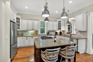 Una cocina con armarios blancos, una isla central, dos taburetes de bar, iluminación colgante y un refrigerador de acero inoxidable.
