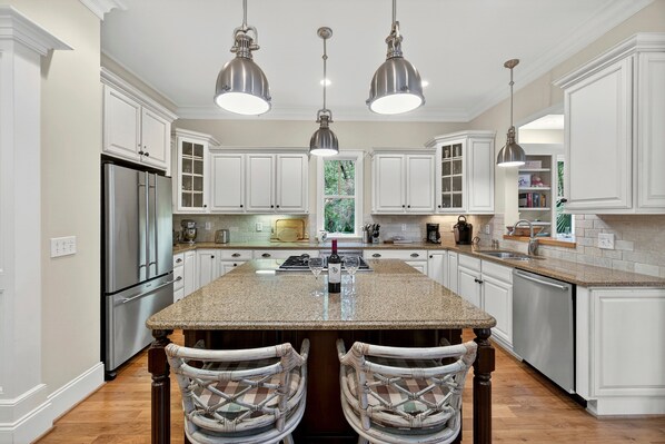 A spacious kitchen with white cabinetry, a central island, pendant lighting, and a view of greenery through the window. - Serene & Private Kiawah Retreat with Private Pool (Kiawah Island)