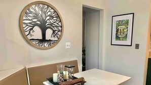 Un salon moderne avec un canapé blanc, une table à manger agrémentée d’un plateau de bougies et d’objets décoratifs, une œuvre d’art représentant un arbre accrochée au mur, et un tableau encadré représentant une maison, également accroché au mur.