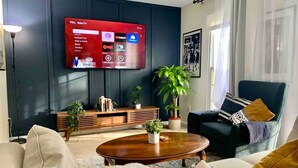Un salon moderne avec un téléviseur à écran plat fixé au mur, un canapé de couleur foncée, une table basse en bois et plusieurs plantes en pot.