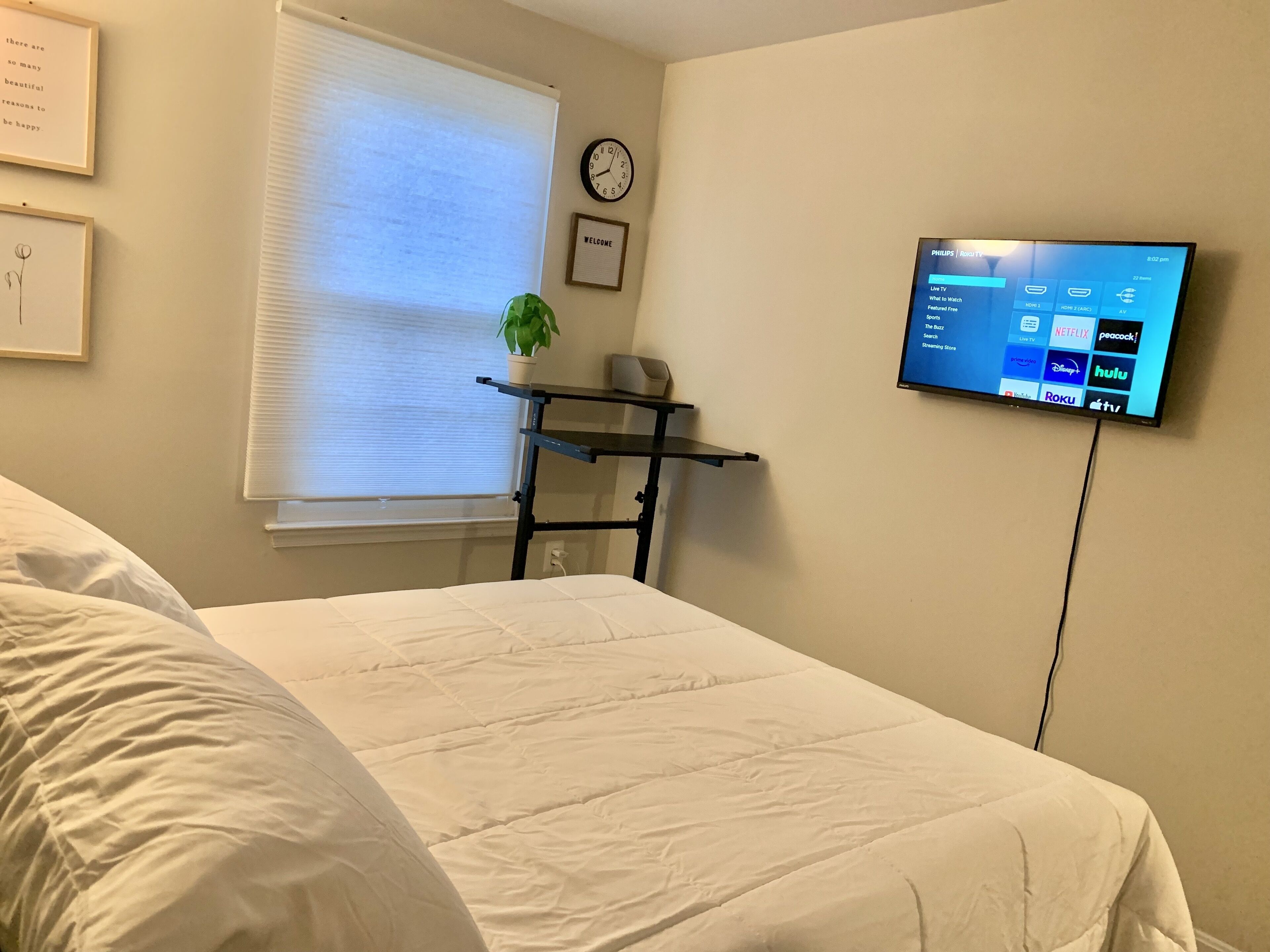A hotel room with a bed, a desk with a plant, a wall clock, and a TV displaying streaming services.