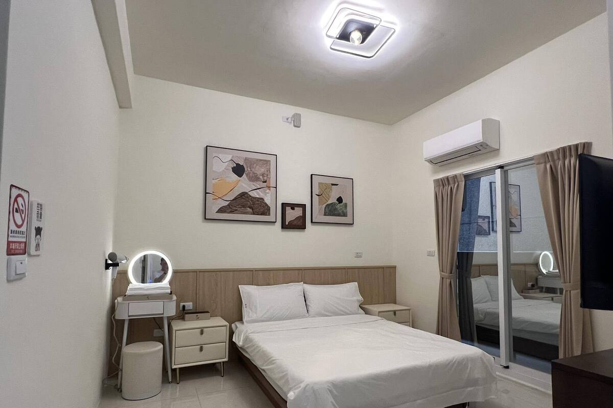Habitación de hotel con cama, mesitas de noche, espejo, decoración en la pared, unidad de aire acondicionado y puerta de cristal corredera.