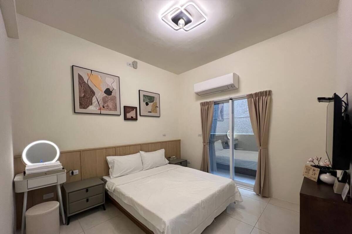 Una habitación de hotel con una cama, una mesita de noche con espejo, un escritorio con computadora, aire acondicionado y una ventana con cortinas.