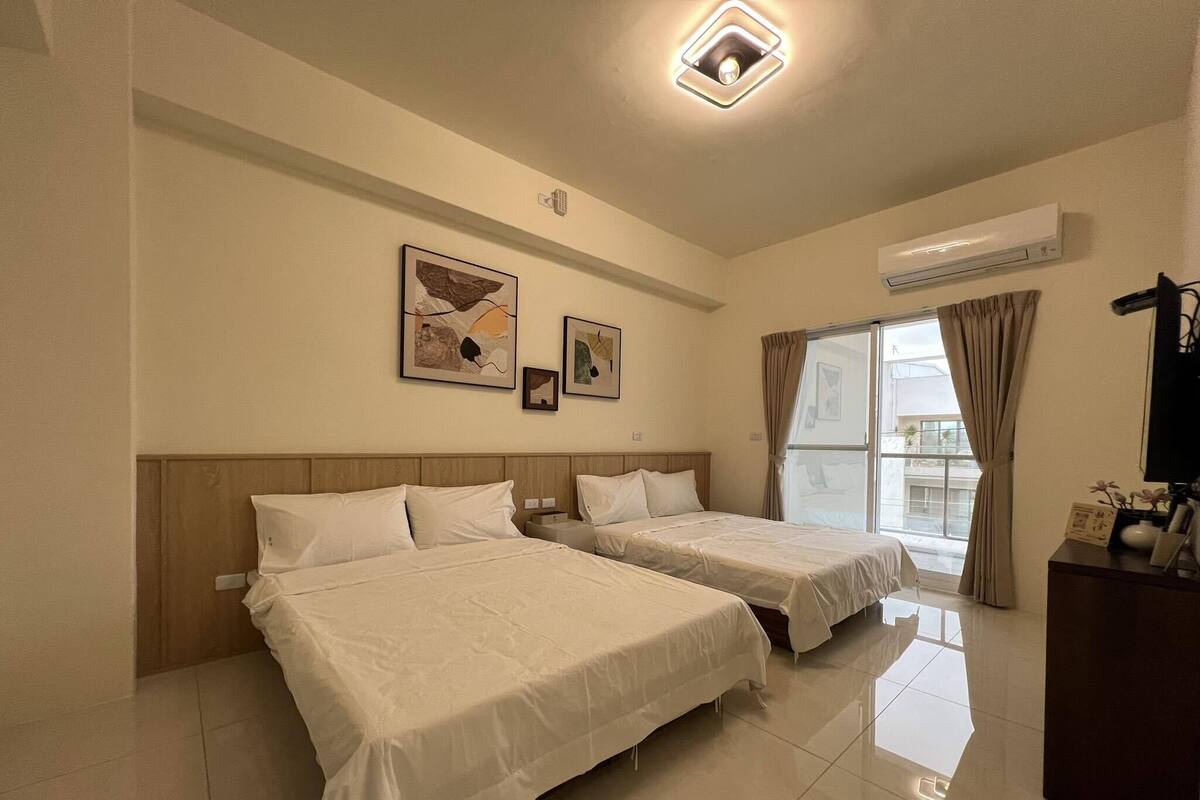 Habitación de hotel con dos camas, una ventana con cortinas, una lámpara de techo y obras de arte colgadas en la pared.