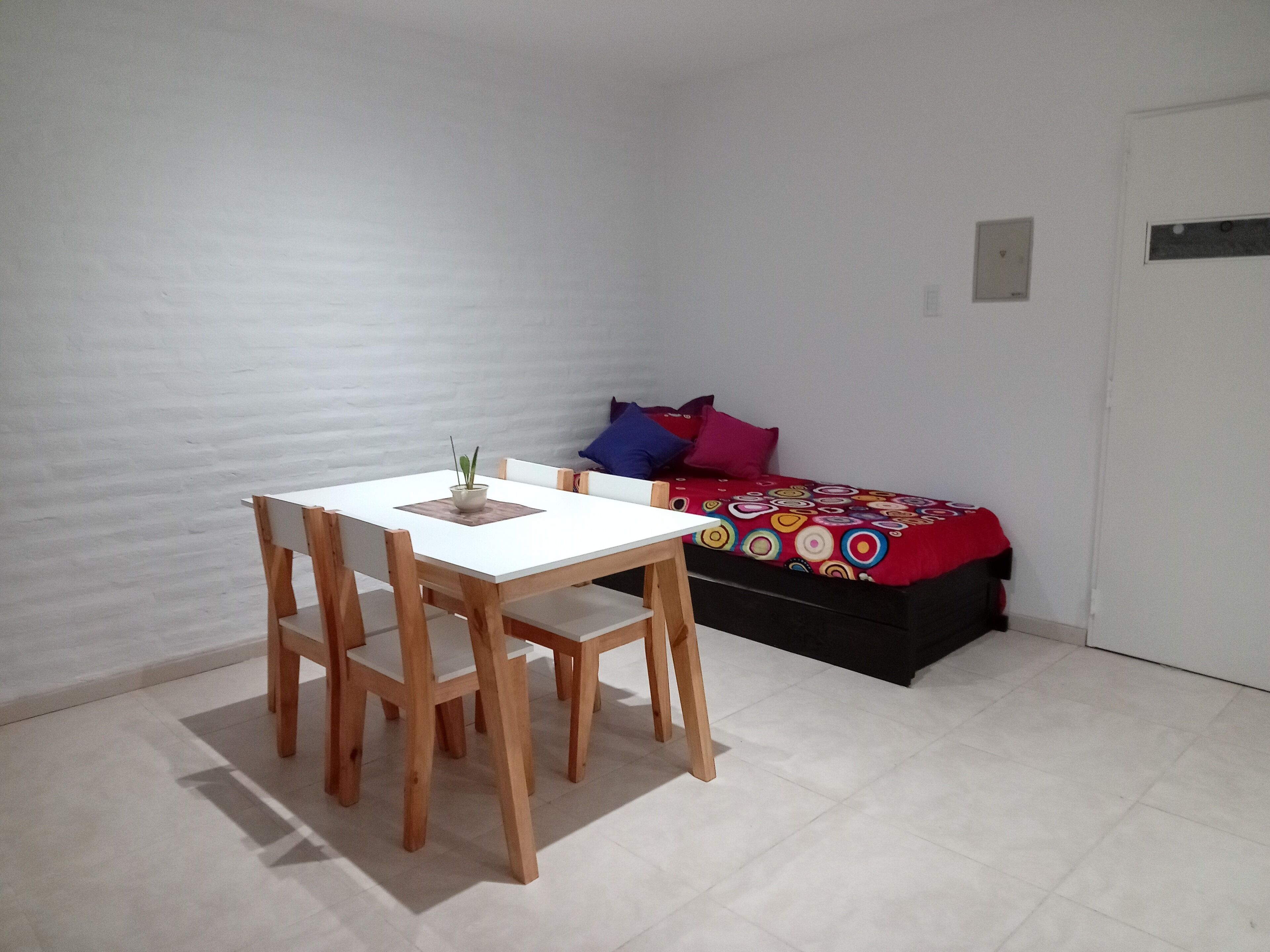 A small room with a white table and wooden chairs, a bed with colorful bedding, and a white wall.