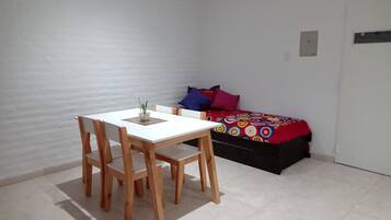 A small room with a white table and wooden chairs, a bed with colorful bedding, and a white wall.