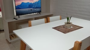 A modern dining area with a white table, wooden chairs, and a television on a shelf displaying a scenic mountain landscape. - Consorcio General Acha ,apartamentos (San Juan)