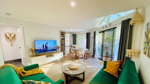 Un moderno salón con sofás verdes, un televisor de pantalla plana, una zona de comedor y un balcón con vistas.